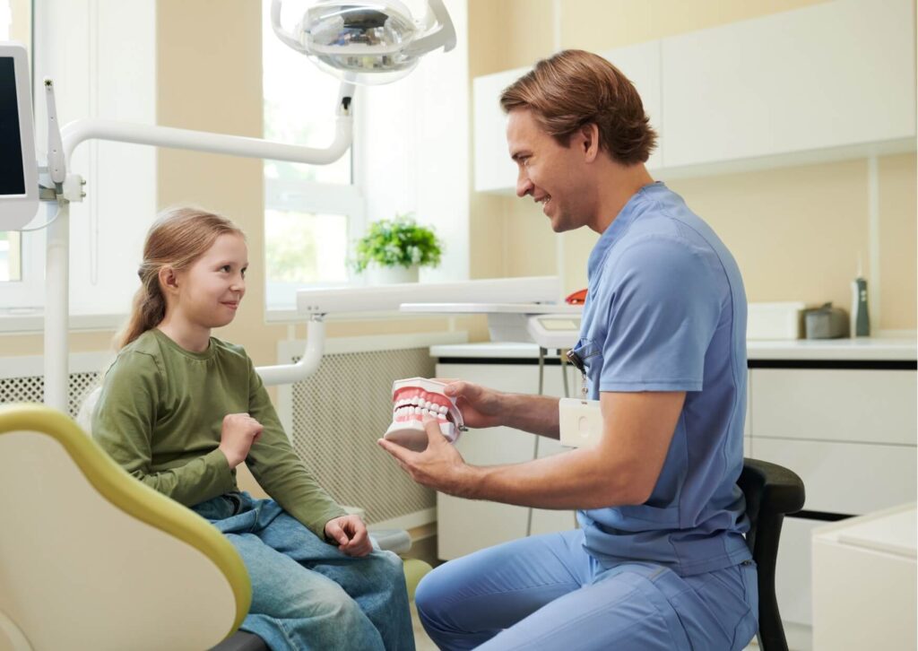 Dentist explaining oral health with a tooth model during a pediatric consultation, helping parents understand when to take their child to the dentist for their first visit.